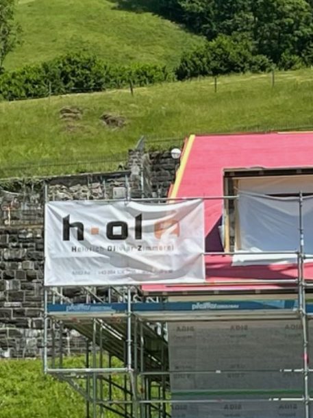 Umbau in Schoppernau, Bregenzerwald neben Kirche in Schoppernau eingerüstetes Haus mit Banner von Heinrich Olivier Zimmerei, Umbau und Sanierung